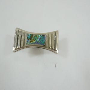 Könnte beinhalten: Silberfarbene Metallbrosche in geschwungener Fliegenform. Die Mitte zeigt eine schillernde Abalone-Einlage mit Grün-, Blau- und Goldtönen. Die Seiten haben vertikale Rippendetails.