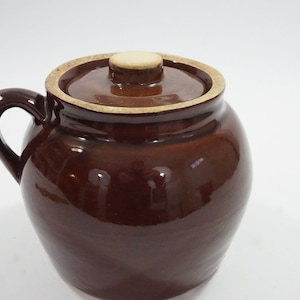 May include: A brown ceramic jar with a lid and handle. The jar has a glossy finish and a light-colored rim. The lid has a small, round knob. This jar is suitable for kitchen storage.