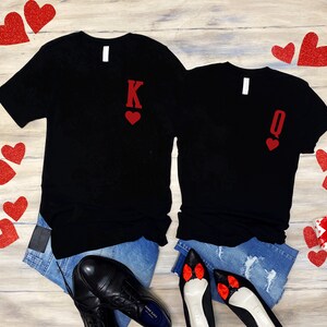 Puede incluir: Dos camisetas negras con corazones rojos de purpurina y las letras "K" y "Q" en ellas. Las camisetas est&aacute;n colocadas sobre una superficie de madera con corazones rojos esparcidos a su alrededor.