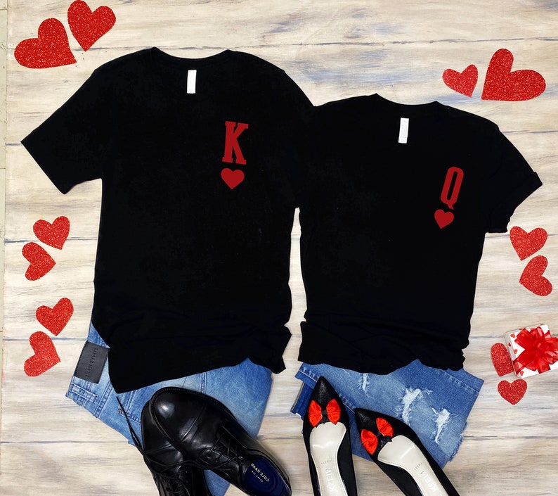Puede incluir: Dos camisetas negras con corazones rojos de purpurina y las letras "K" y "Q" en ellas. Las camisetas est&aacute;n colocadas sobre una superficie de madera con corazones rojos esparcidos a su alrededor.