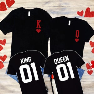 Puede incluir: Dos camisetas negras con corazones rojos y las palabras "King 01" y "Queen 01" impresas en ellas. Las camisetas est&aacute;n sobre una superficie de madera con corazones rojos esparcidos a su alrededor.