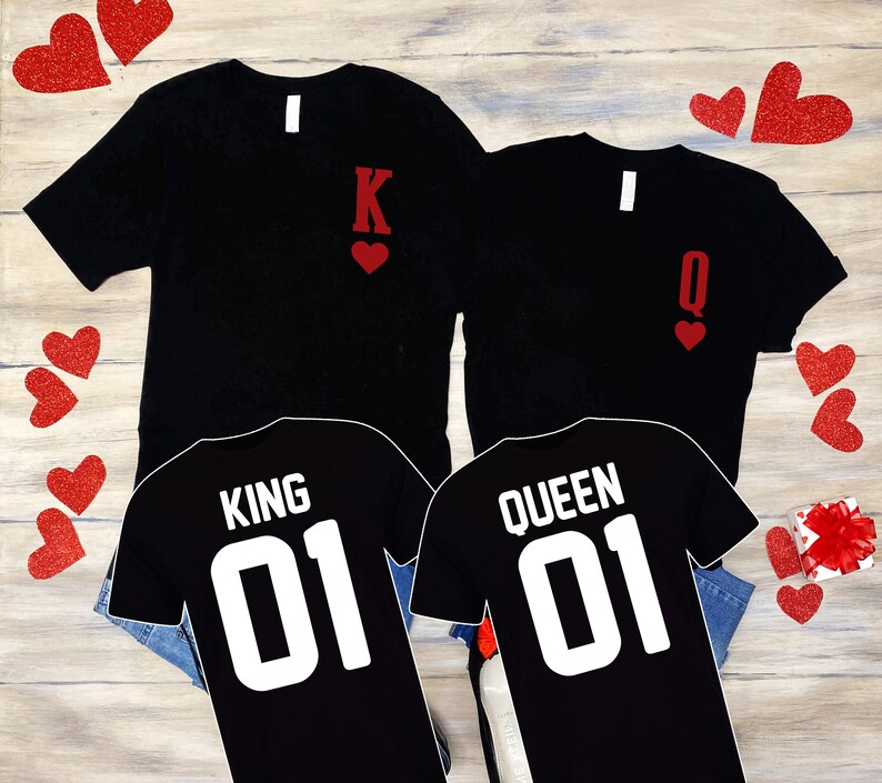 Puede incluir: Dos camisetas negras con corazones rojos y las palabras "King 01" y "Queen 01" impresas en ellas. Las camisetas est&aacute;n sobre una superficie de madera con corazones rojos esparcidos a su alrededor.