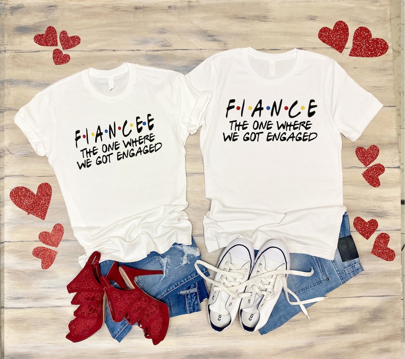 K&ouml;nnte beinhalten: Zwei wei&szlig;e T-Shirts mit dem Text "FIANCEE THE ONE WHERE WE GOT ENGAGED" und "FIANCE THE ONE WHERE WE GOT ENGAGED" in schwarzer Schrift. Die Shirts liegen auf einer Holzoberfl&auml;che mit roten Herzen, einer blauen Jeans und wei&szlig;en Turnschuhen.
