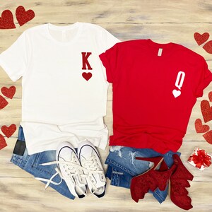 Puede incluir: Una camiseta blanca con una 'K' roja y un coraz&oacute;n, y una camiseta roja con una 'Q' blanca y un coraz&oacute;n. Ambas camisetas est&aacute;n sobre una superficie de madera con corazones rojos esparcidos a su alrededor.