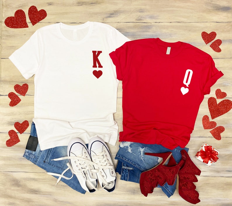 Puede incluir: Una camiseta blanca con una 'K' roja y un coraz&oacute;n, y una camiseta roja con una 'Q' blanca y un coraz&oacute;n. Ambas camisetas est&aacute;n sobre una superficie de madera con corazones rojos esparcidos a su alrededor.