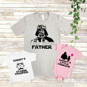 May include: Three t-shirts with Star Wars themed designs. The first shirt is grey with a black image of Darth Vader and the text "I am your father". The second shirt is white with a black image of a Stormtrooper and the text "Daddy's Storm Pooper". The third shirt is pink with a black image of Darth Vader and a child and the text "Daddy's Little Princess".