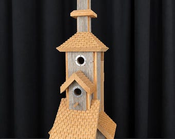 Può includere: Una casetta per uccelli in legno a più livelli con rivestimento in legno grigio usurato e tetti in scandole marrone chiaro. La struttura presenta tre fori d'ingresso circolari con bordo in metallo, ciascuno dotato di un piccolo posatoio in legno.