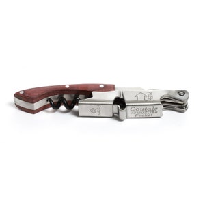 May include: A silver and wood wine opener with a wooden handle and a silver lever. The opener has a corkscrew, a foil cutter, and a bottle stopper. The text "Coutale Sommelier Pocket" is engraved on the opener.