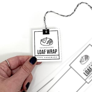 Könnte beinhalten: Ein schwarz-weißes Etikett mit dem Text "Sourdough Loaf Wrap 100% Handmade" und einer Grafik eines Laibes Brot. Das Etikett ist an einer weißen Schnur befestigt.