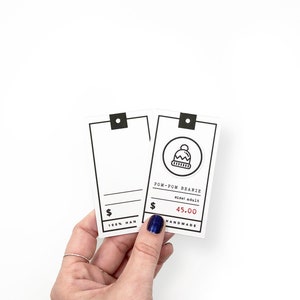 May include: Two white price tags with black borders and text. The top tag has a black and white illustration of a beanie with a pom-pom. The text reads "Pom-Pom Beanie", "size: adult", "$45.00", and "Handmade". The bottom tag has the text "100% Han"