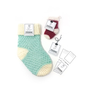 Puede incluir: Dos calcetines de Navidad de ganchillo, uno grande y otro pequeño. El calcetín grande es azul claro y crema, el calcetín pequeño es rojo y crema. Ambos calcetines tienen una etiqueta blanca con texto negro que dice "Holiday Stocking".