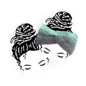 May include: A grey and teal knitted headband with a twist design, displayed on a white heart-shaped card with a black and white illustration of a woman's face and hair. The headband is a soft, textured accessory.