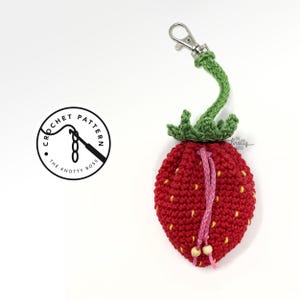 May include: A red crocheted strawberry-shaped pouch with a green stem and leaves, a pink drawstring, and a silver clasp. The pouch has yellow seed details and two wooden beads. The logo for "The Knotty Boss" is in the upper left corner.
