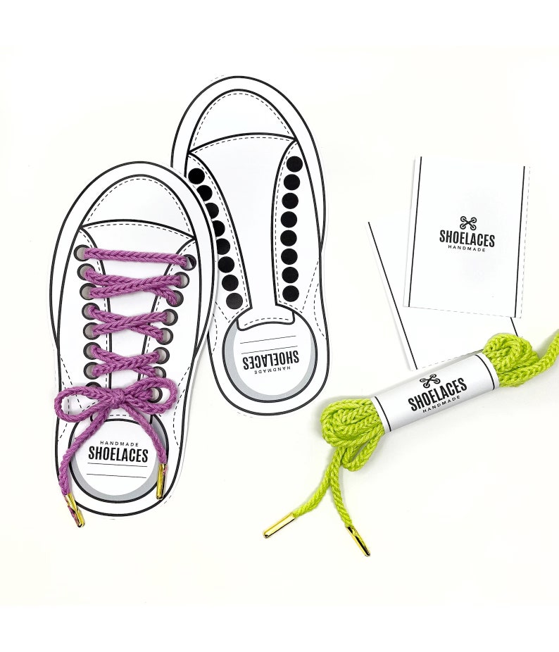 May include: A pair of white canvas shoes with black dots, one with purple shoelaces tied in a bow, and the other with a shoelace loop. A green shoelace with gold aglets is wrapped around a white label with the text "Shoelaces Handmade".