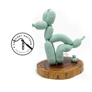 Puede incluir: Escultura de perro globo de ganchillo en color verde claro, exhibida sobre una base de madera. La escultura está hecha de hilo y presenta un diseño detallado. El logotipo "Crochet Pattern The Knotty Boss" es visible.