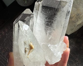 May include: A clear quartz crystal cluster featuring multiple pointed terminations with translucent facets and a small patch of light brown mineral inclusion on one of the primary crystal faces, showcasing natural geometric growth patterns.