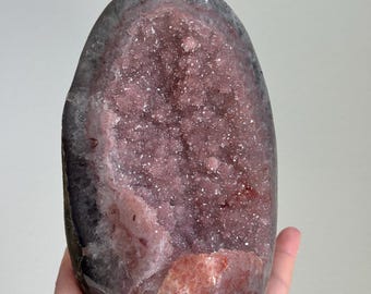 Op de afbeelding: Een gepolijste ovale geode met een diep, kristallijn interieur in tinten van oudroze en mauve. De buitenrand van de steen is een gladde, donkergrijze minerale behuizing die de getextureerde, sprankelende roze kwartskristallen omlijst.