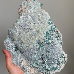 Op de afbeelding: Een groot, onregelmatig mineraal exemplaar met een getextureerd oppervlak met een mix van diep bosgroen, bleek muntgroen en lichtgrijze kristallijne afzettingen. De steen heeft een ruwe, grillige rand en vertoont diverse minerale insluitingen.