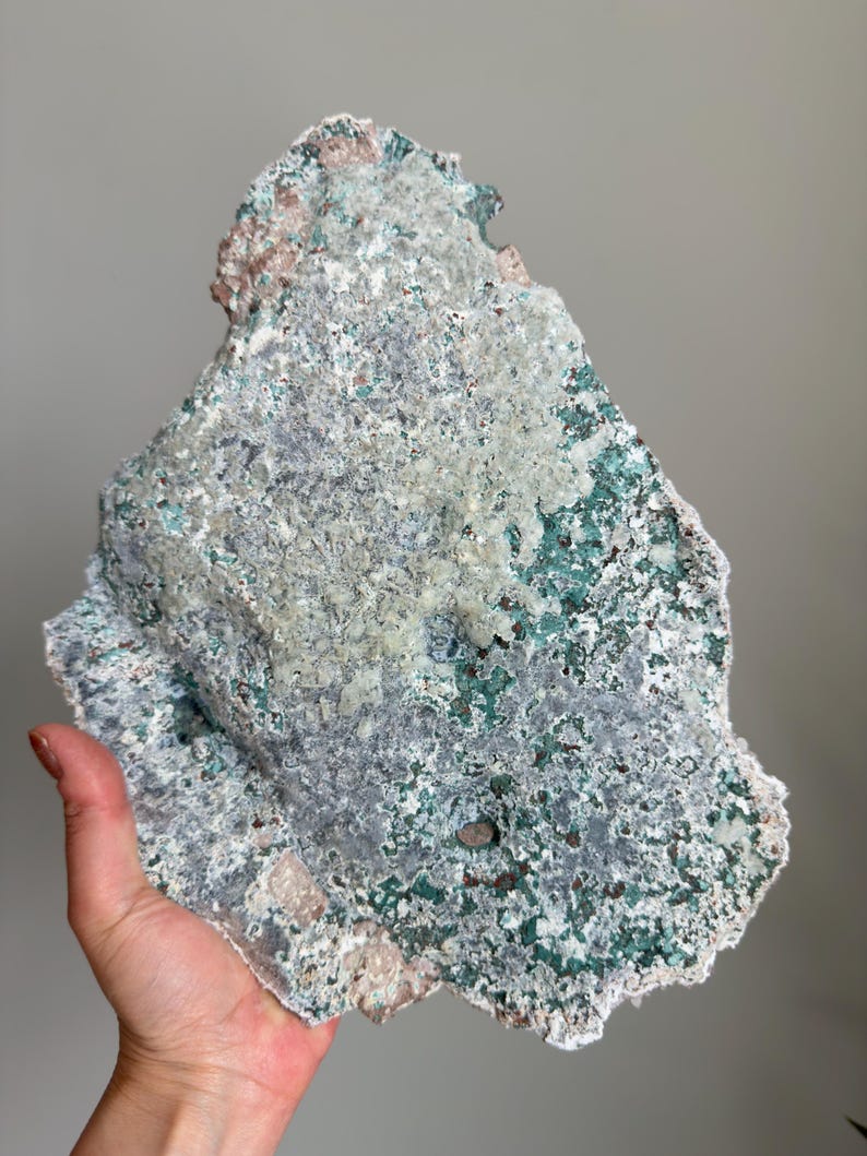 Op de afbeelding: Een groot, onregelmatig mineraal exemplaar met een getextureerd oppervlak met een mix van diep bosgroen, bleek muntgroen en lichtgrijze kristallijne afzettingen. De steen heeft een ruwe, grillige rand en vertoont diverse minerale insluitingen.