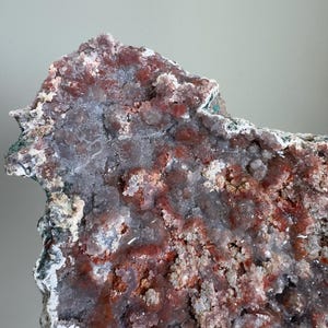 Op de afbeelding: Een groot mineraalexemplaar met een cluster van kristallijne formaties in tinten van diep roodbruin, doorschijnend grijs en wit. Het oppervlak is getextureerd met kleine, grillige kristalpunten en ruwe, onregelmatige randen overal.
