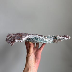 Op de afbeelding: Een plat, breed mineraal exemplaar met een bovenoppervlak bedekt met kleine, getextureerde, oudroze kristallijne formaties. De onderkant en randen vertonen een ruwe, poreuze textuur met gevlekte groene, witte en grijze mineraalafzettingen.