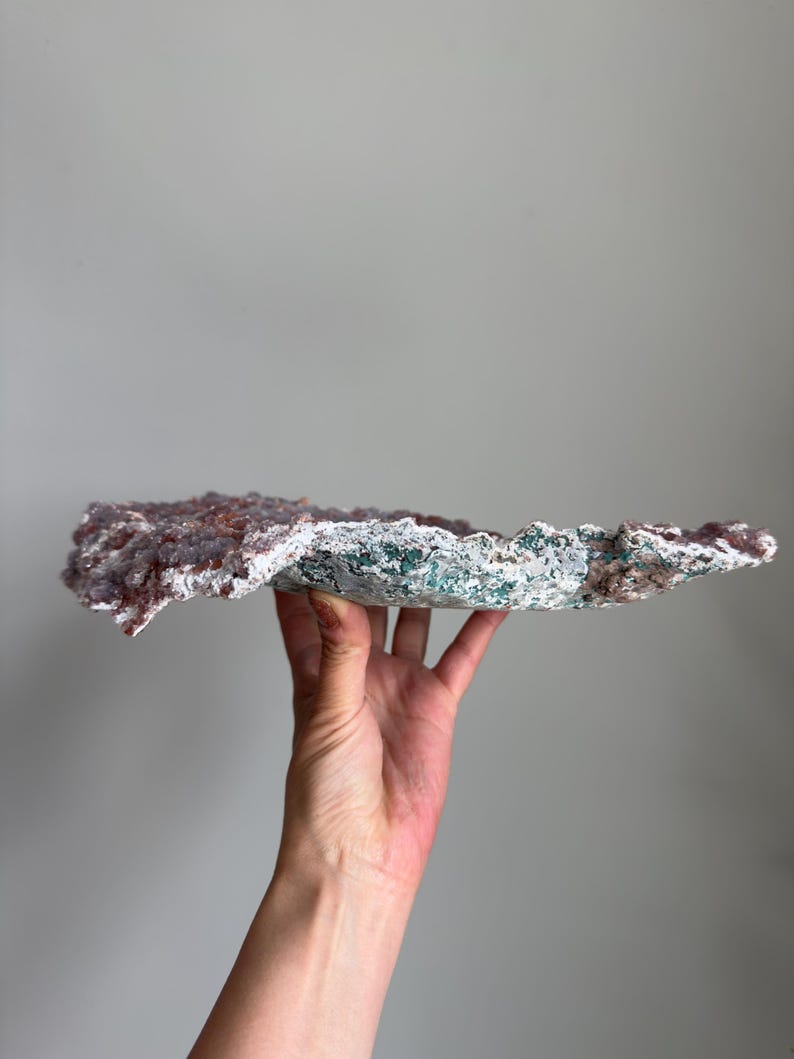 Op de afbeelding: Een plat, breed mineraal exemplaar met een bovenoppervlak bedekt met kleine, getextureerde, oudroze kristallijne formaties. De onderkant en randen vertonen een ruwe, poreuze textuur met gevlekte groene, witte en grijze mineraalafzettingen.