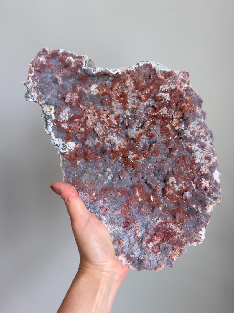 Op de afbeelding: Een groot, plat mineraalexemplaar met een textuurrijk oppervlak bedekt met clusters van kleine, kristallijne formaties. Het stuk vertoont een gevlekt kleurenpalet van diep roodbruin en gedempt lavendelgrijs, met een ruwe, onregelmatige rand.