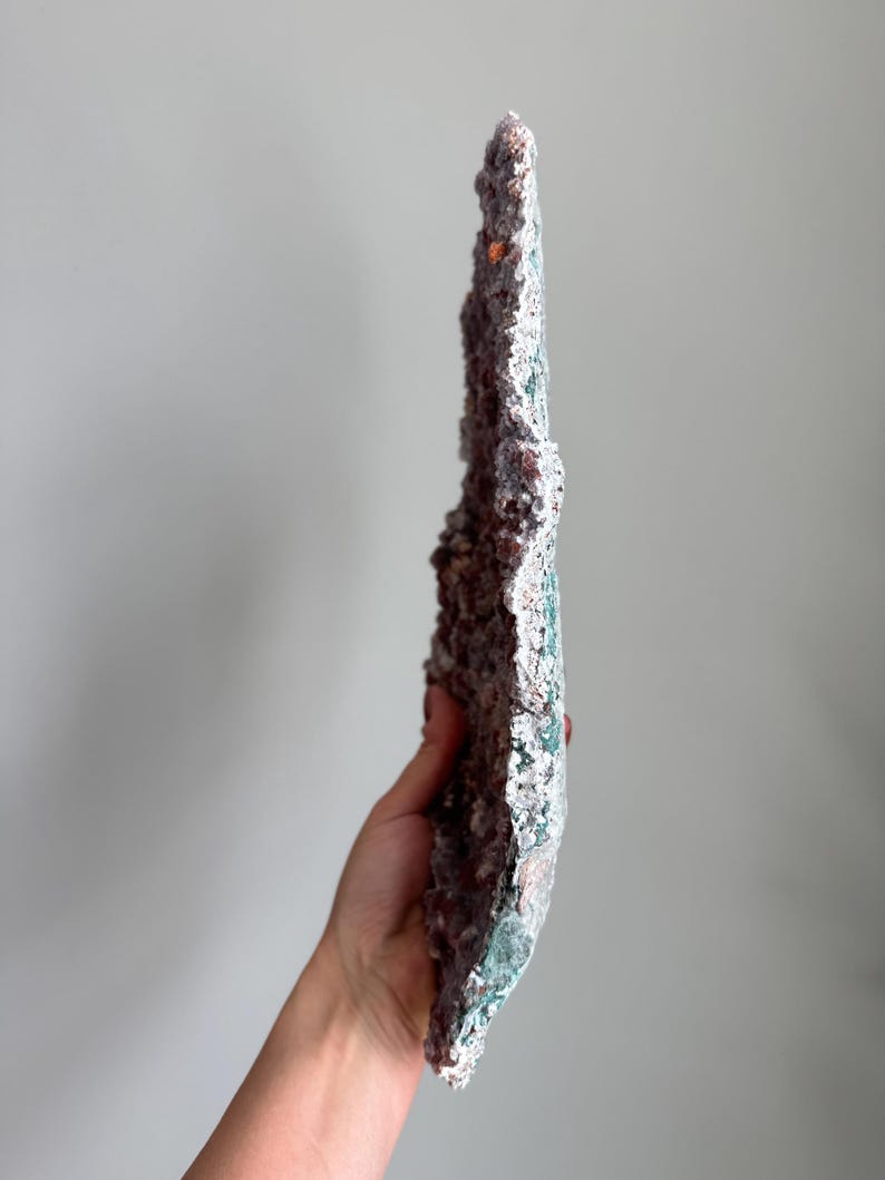 Op de afbeelding: Een lang, dun, verticaal mineraalexemplaar met een ruwe, kristallijne textuur. Het oppervlak vertoont een mix van diepe roodbruine en aardse tinten, met vlekken van levendige blauwgroene mineraalafzettingen langs de buitenste, grillige randen.