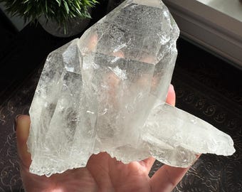 May include: A large, clear quartz crystal cluster featuring multiple pointed terminations and a translucent, multifaceted structure. The mineral specimen displays natural internal fractures and inclusions, with a jagged base and sharp, geometric edges.