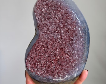 Op de afbeelding: Een niervormig geode-exemplaar met een diep roodpaars kristallijn interieur met een sprankelend, getextureerd oppervlak. De buitenrand van de steen is omhuld door een gladde, donkergrijze minerale laag die een duidelijk contrast vormt met het midden.