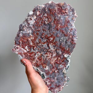 May include: A large, flat mineral specimen featuring a textured surface with a mix of deep reddish-brown and muted purple crystalline formations. The irregular, jagged edges define the shape of this geological piece, showcasing a dense cluster of small, sparkling mineral growths across its entire front-facing surface.