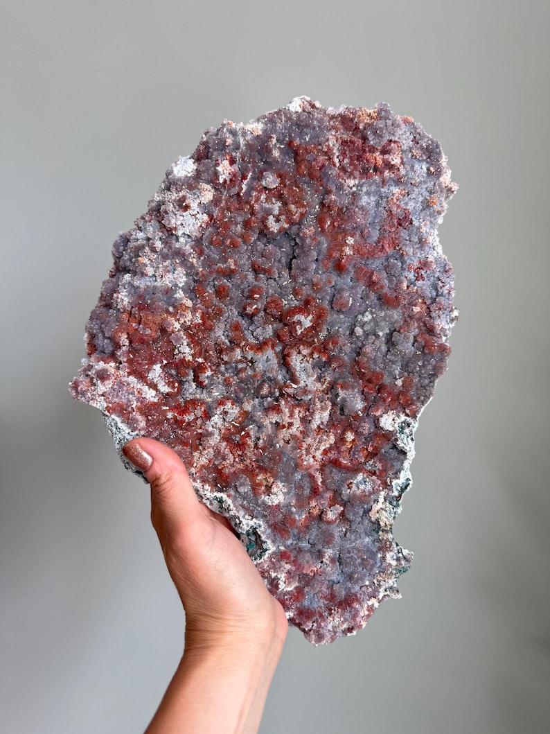 Op de afbeelding: Een groot, plat mineraalexemplaar met een getextureerd oppervlak met een mix van dieproodbruine en gedempte paarse kristallijne formaties. De onregelmatige, grillige randen bepalen de vorm van dit geologische stuk, met een dichte cluster van kleine, sprankelende mineraalgroei over het gehele vooroppervlak.