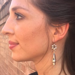Puede incluir: Pendientes colgantes plateados con un diseño floral en la parte superior y forma de lágrima en la parte inferior. Los pendientes los lleva una persona con cabello oscuro y una parte superior negra. El fondo es una pared de ladrillos.