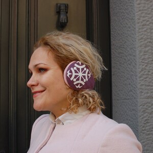 May include: A woman wearing a pink jacket with a purple ear warmer featuring a white snowflake design.