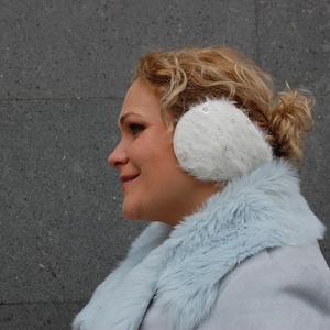 May include: A woman wearing a light blue faux fur coat with white earmuffs. The earmuffs are made of fluffy white material and have a small white button on the side.
