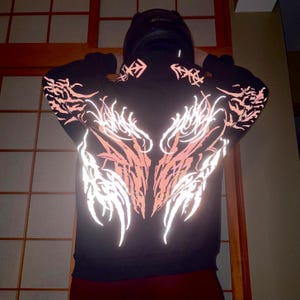 Può includere: Felpa con cappuccio nera con design tribale riflettente rosa e bianco sulla schiena e sulle maniche. Il design si illumina con la luce. Indossata da una persona con casco nero.