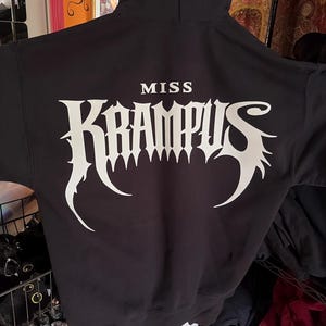 Könnte beinhalten: Schwarzer Kapuzenpullover mit dem weißen Schriftzug "MISS KRAMPUS" in gotischer Schrift. Der Pullover hat eine Kapuze mit Kordelzug und eine Vordertasche. Das Design ist auf der Rückseite des Kleidungsstücks aufgedruckt.
