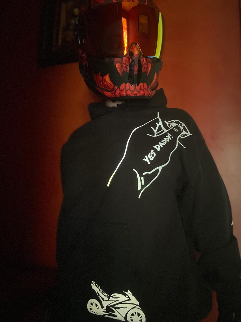 May include: Black hoodie with the text "YES DADDY!" and a motorcycle graphic. The hoodie is from "CRTY'S CLOSET". The person is wearing a motorcycle helmet with a red and black design.