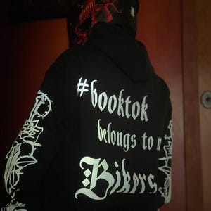 May include: Black hoodie with white text that reads "#booktok belongs to Bikers." The sleeves have white graphic designs. A red and black helmet is visible.