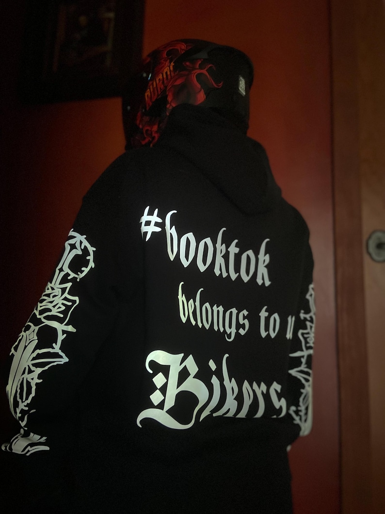 May include: Black hoodie with white text that reads "#booktok belongs to Bikers." The sleeves have white graphic designs. A red and black helmet is visible.