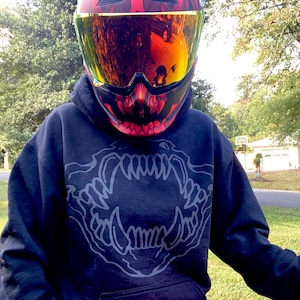 May include: A motorcycle helmet with a red and black design, featuring a gold visor. The helmet has a graphic of a skull on the front. The person is wearing a black hoodie with a grey jaw graphic.