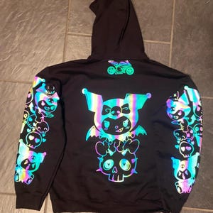 Könnte beinhalten: Schwarzer Kapuzenpullover mit einem reflektierenden, farbenfrohen Design eines Zeichentrickcharakters mit Totenköpfen und einem Motorrad. Das Design befindet sich auf dem Rücken und den Ärmeln. Der Kapuzenpullover hat eine Kapuze.