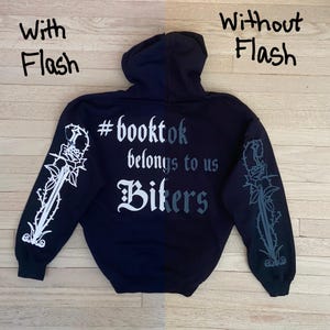 May include: A navy blue hoodie with the text "#booktok belongs to us Bikers" on the back. The sleeves feature a rose and dagger design. The image is shown with and without flash.