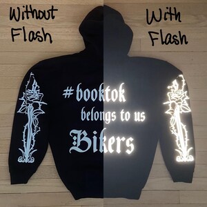 May include: Black hoodie with the text "#booktok belongs to us Bikers" in white, along with a rose and thorn design on the sleeves. The hoodie has a hood and long sleeves. The text and design are reflective.