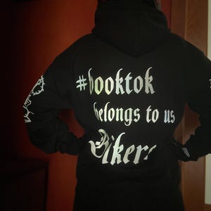 May include: Black hoodie with the text "#booktok belongs to us Bikers" in a reflective, silver font. The hoodie has a hood and long sleeves. The person is wearing a black helmet with red flames.