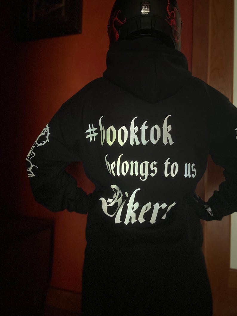 May include: Black hoodie with the text "#booktok belongs to us Bikers" in a reflective, silver font. The hoodie has a hood and long sleeves. The person is wearing a black helmet with red flames.