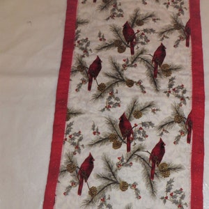 May include: Red cardinal birds on a white background with pine branches and red berries. The table runner has a red border with white polka dots.