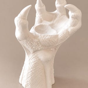 Pode incluir: Uma escultura branca mate em forma de garra ou pata grande e texturizada. A superfície apresenta padrões intrincados em relevo semelhantes a escamas e sulcos profundos, com quatro garras proeminentes, curvas e pontiagudas que se elevam da base.