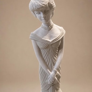 Può includere: Una statuetta bianco opaco raffigurante una persona con capelli corti, che indossa un abito con spalle scoperte, corpetto plissettato e gonna svasata. La figura presenta una collana girocollo dettagliata e un bracciale, con le mani giunte davanti.