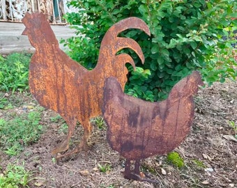 May include: Two rusty metal silhouettes of a rooster and a hen. The rooster is standing upright with its tail feathers spread out. The hen is standing next to the rooster with its head turned to the side. The silhouettes are on a bed of brown mulch.